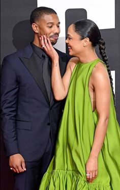 a man and woman standing next to each other in front of a black wall wearing green dresses