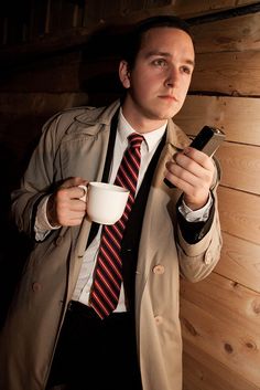 a man in a trench coat holding a cup and cell phone while standing next to a wooden wall