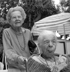 an older woman sitting in a chair next to another old woman with glasses on her head