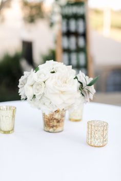 white flowers in vases sitting on top of a table next to two gold candles