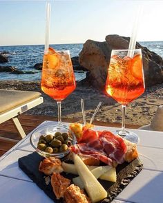 two glasses of wine and some food on a table near the water with an ocean in the background