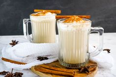 two mugs filled with white liquid sitting on top of a wooden board next to cinnamon sticks