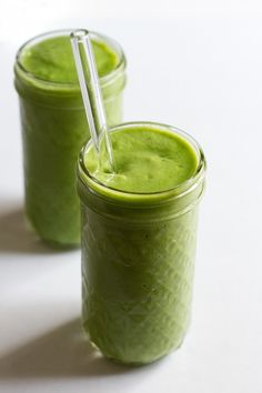 two mason jars filled with green smoothies on a white surface, one has a straw in it and the other is empty