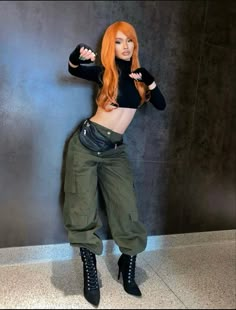 a woman with long red hair wearing green pants and black boots is posing for the camera