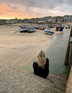 a woman sitting on the edge of a pier looking at boats in the water and buildings