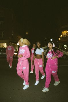 three women in pink tracksuits are walking down the street at night with their arms around each other