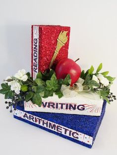 a red apple sitting on top of two books with greenery and flowers around it