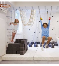 two children are playing in a room with climbing ropes and boxes on the floor, while one child is jumping