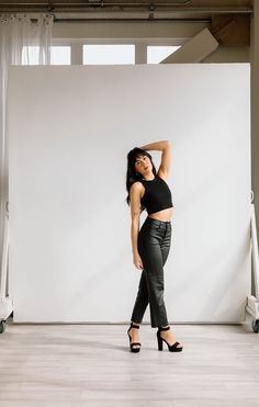 a woman standing in front of a white wall with her arms behind her head and wearing high heels