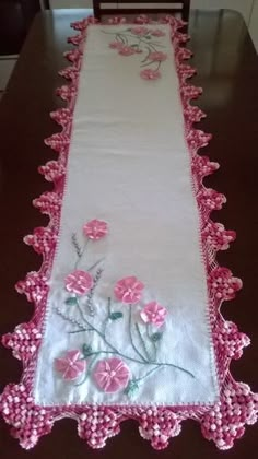 a table runner with pink flowers on white and pink trimmings, sitting on top of a wooden table