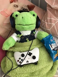 a stuffed frog sitting on top of a bed with headphones and a video game controller