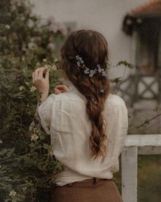 a woman with long hair wearing a white blouse and brown skirt standing in front of flowers