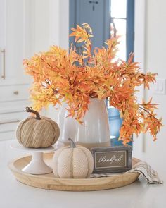 a white vase filled with orange and yellow leaves on top of a wooden platter