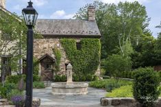 an old stone house with a fountain in the front yard, surrounded by greenery