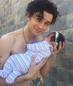 a shirtless man holding a baby in his arms while standing next to a stone wall