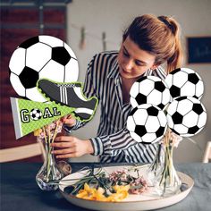 a woman arranging vases with flowers and soccer balls in them on a table top