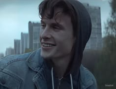 a young man wearing a denim jacket and hoodie looks off into the distance in front of a cityscape