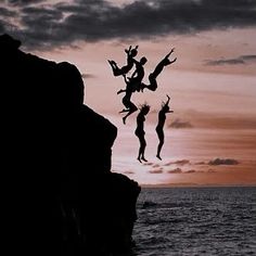 three people jumping off rocks into the ocean at sunset or dawn with their arms in the air