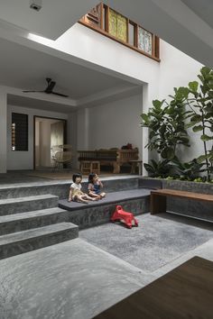 two children are sitting on some steps in a house with large plants and stairs leading up to the second floor