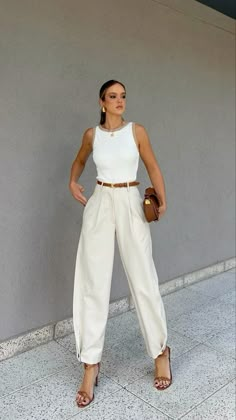 a woman standing in front of a gray wall wearing white pants and a white top