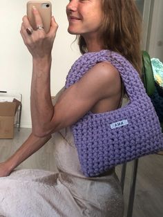 a woman sitting down holding a cell phone and wearing a purple crocheted bag