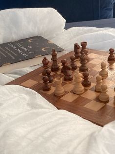 a wooden chess set on top of a bed next to a book and some pillows