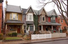 a row of houses on a street with trees in the foreground and a white picket fence