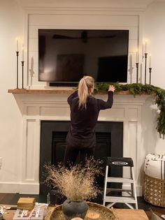 a woman standing in front of a flat screen tv above a fireplace with candles on it