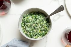 a bowl filled with spinach on top of a white table next to glasses of wine