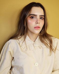 a woman with long hair wearing a white shirt and black pants, standing against a yellow wall