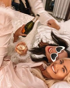 two beautiful women laying on top of a bed next to each other holding wine glasses