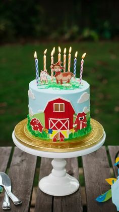 a birthday cake decorated with farm animals and lit candles on a wooden table in front of a grassy field