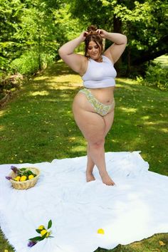 A woman adjusts her hair while posing barefoot on a white blanket in a serene park setting. A wicker basket with lemons and flowers is placed to her side, and sunlight filters through the trees.