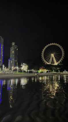 the ferris wheel is lit up at night by the water's edge with buildings in the background