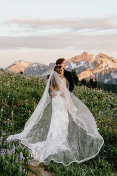 a bride and groom standing on top of a grass covered field with mountains in the background