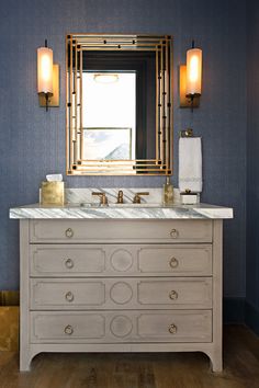 a bathroom vanity with marble top and gold accents on the mirror above it is decorated in blue wallpaper