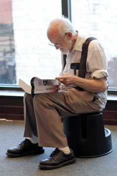 an older man sitting on top of a chair reading a book in front of a window