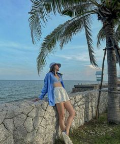 a woman leaning on a stone wall next to the ocean with palm trees in the background