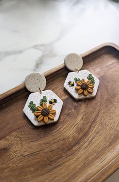 two earrings with sunflowers and leaves on them sitting on a wooden tray next to a marble counter top