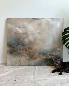 a cat laying on top of a white bed next to a painting and a potted plant