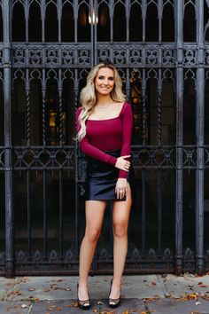 a woman standing in front of an iron gate wearing a red top and black skirt