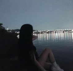 a woman sitting on the edge of a cliff looking out over water at night time