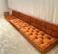 a long orange couch sitting in front of a window with curtains on the windowsill