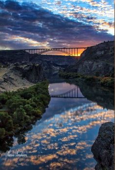 a bridge over a body of water under a sky filled with clouds and trees at sunset