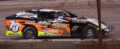 an orange and black race car on the track behind a chain link fence with other cars in the background