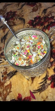 a glass bowl filled with cereal on top of a floral table cloth next to a metal spoon