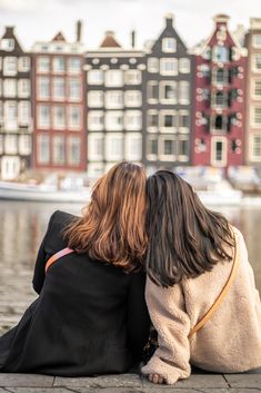 two women sitting on the ground facing each other with buildings in the background and water behind them