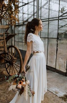 a woman standing in front of a greenhouse holding a bouquet of flowers and wearing a white dress