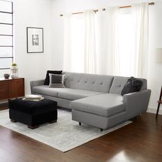 a living room with a couch, ottoman and table in front of two large windows