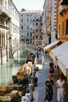 several people walking down a narrow street next to buildings and a canal with boats on it